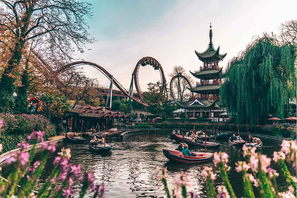 A view of the Tivoli gardens with pink flowers and people riding small boats on a pond in the foreground and a rollercoaster and pagoda-styled building in the background