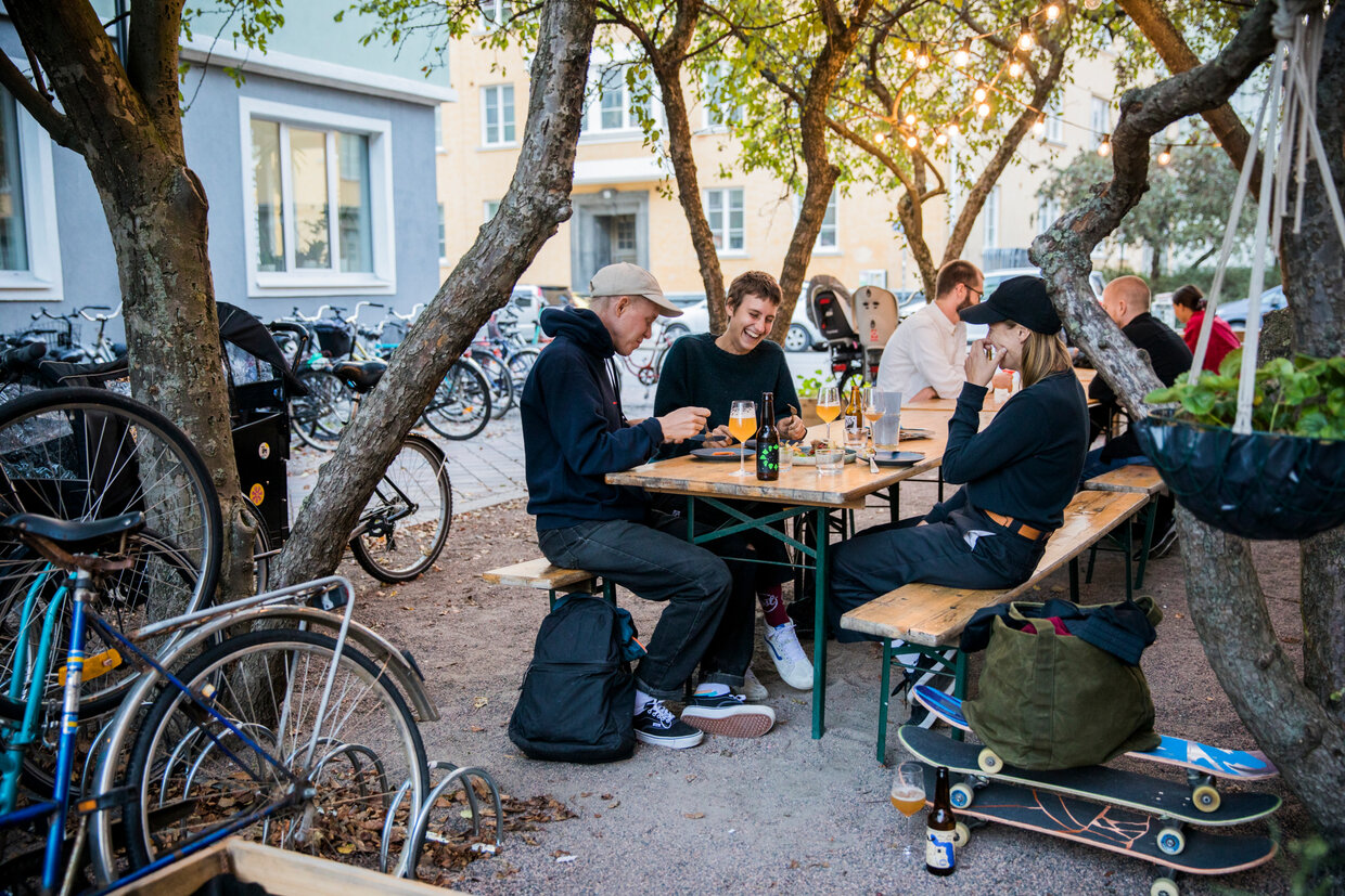 Three young men laughing while enjoying drinks at a table in the outdoor seating area of a café or bar